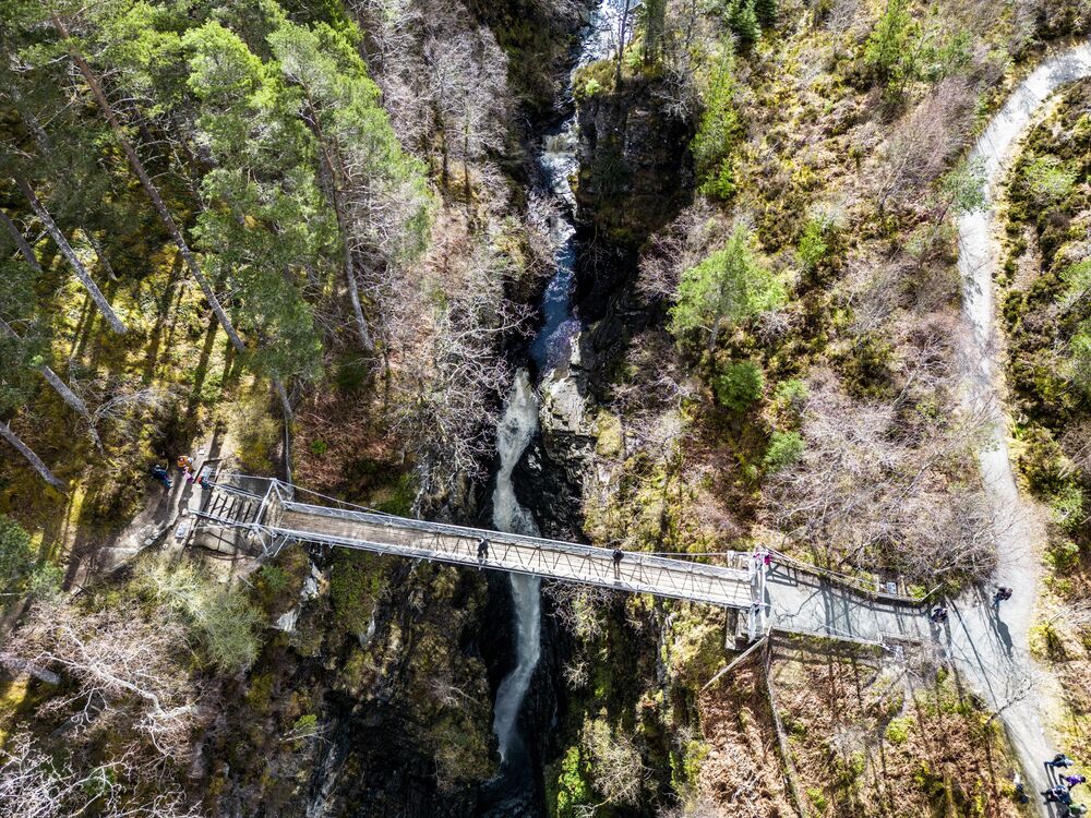 An aerial view of a narrow suspension bridge spanning a deep gorge. A waterfall crashes down the gorge. Woodland surrounds the banks either side.
