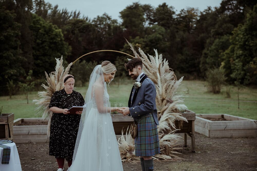 A bride and groom exchange rings during their wedding ceremony. The celebrant stands just behind them, beside a woven willow arch. An orchard is in the background.
