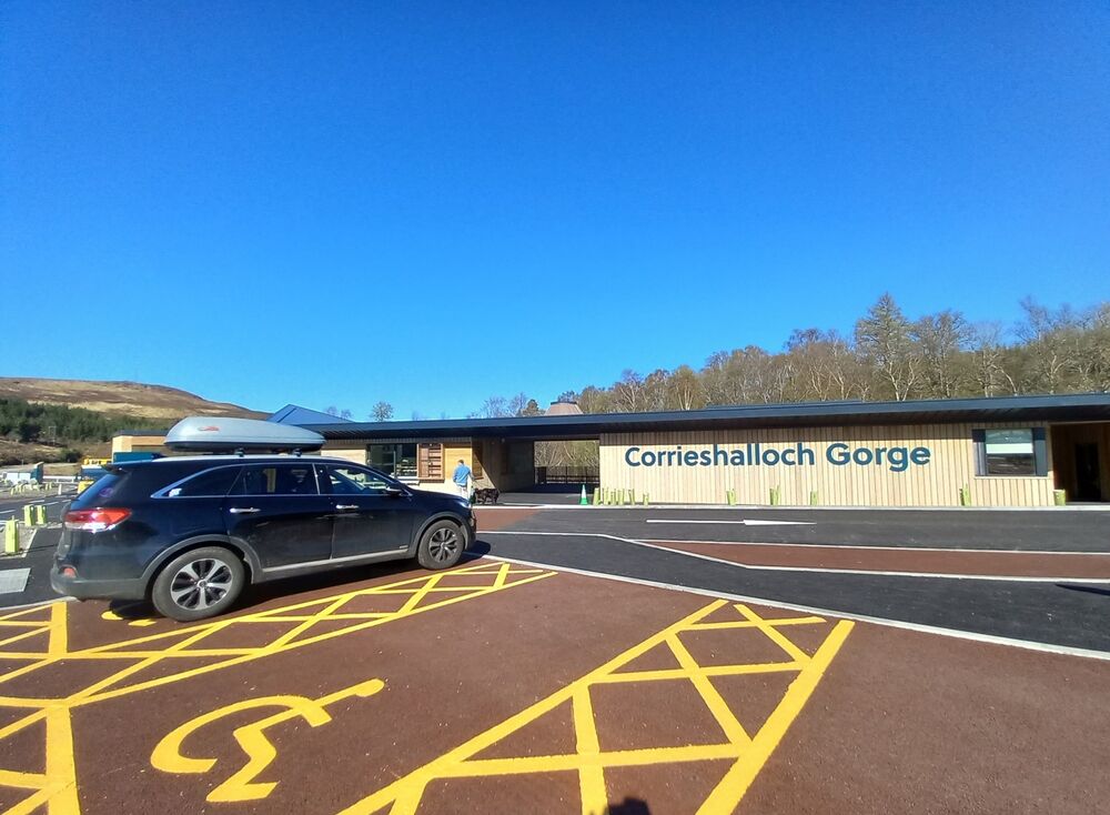 Two parking bays in a car park are clearly designated for disabled users. They are a short distance from a visitor centre. A car is parked in the bay on the left.