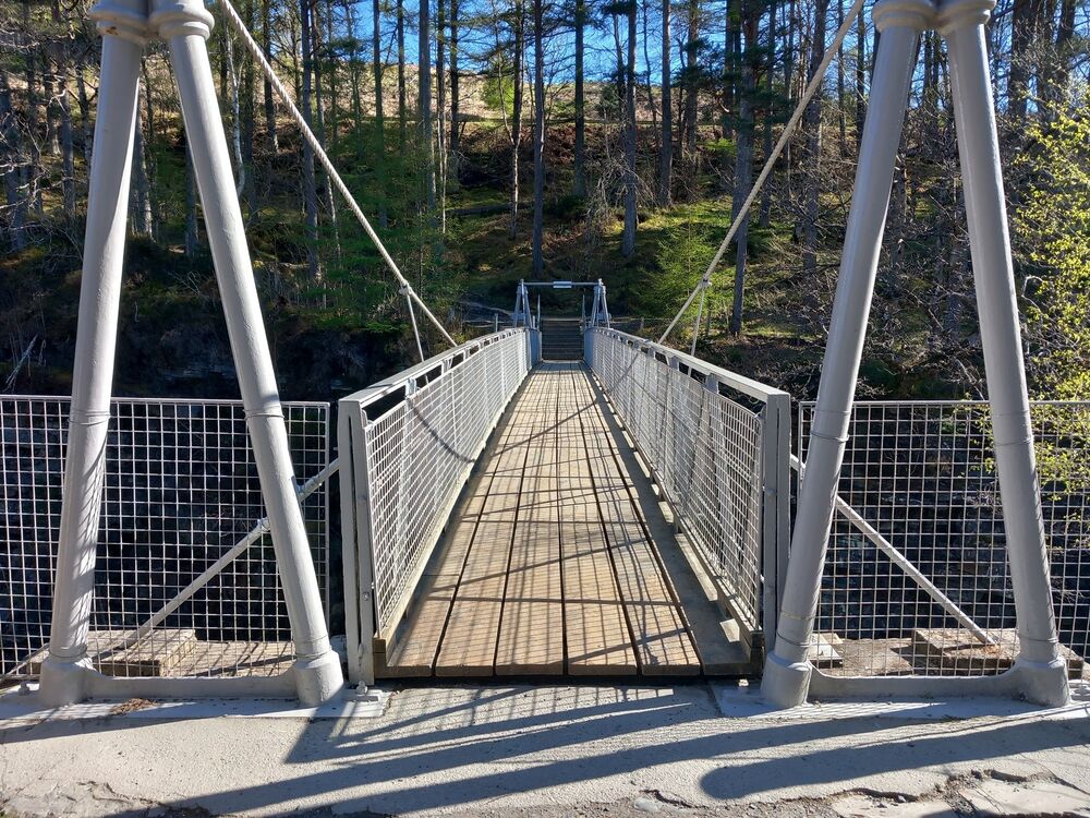 A view looking directly across a narrow suspension bridge for pedestrians. It has metal railings and cables, with wooden boards on the walkway.