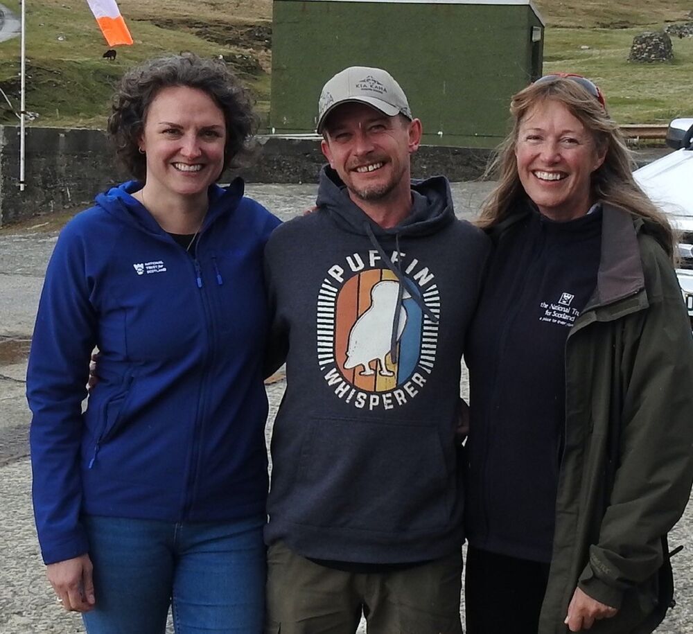 Three people stand in a row, their arms around each other, and smile at the camera. A man wearing a puffin sweater stands in the middle, with a woman either side. Both women are wearing National Trust for Scotland jumpers.