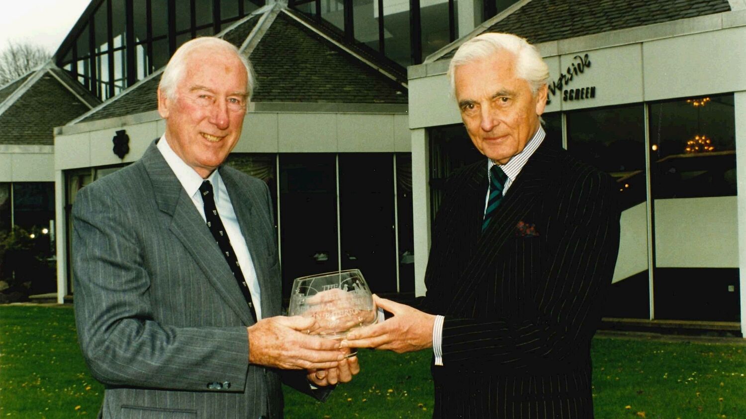 Two older men stand outside a modern building, holding an engraved glass trophy between them.