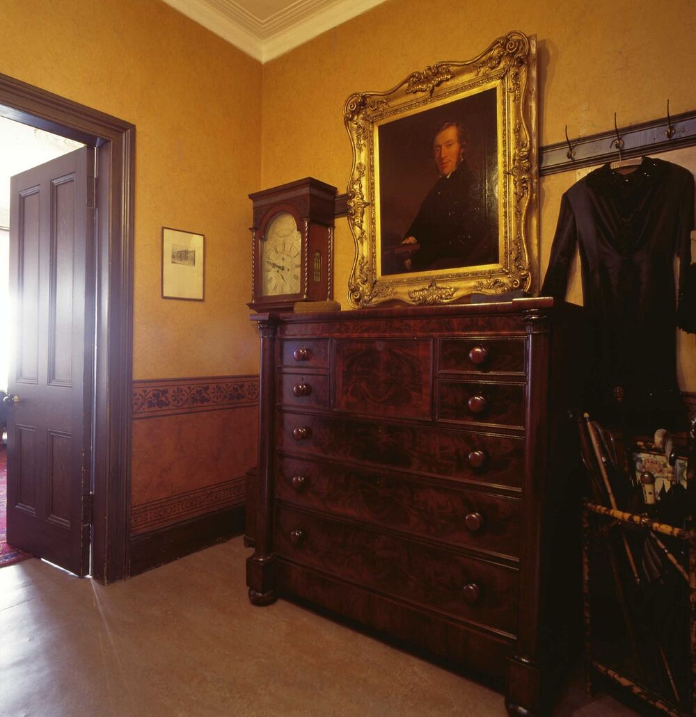 A view of the hall in the Tenement House, showing a yellow wallpaper. A dark wooden dresser stands at the far end next to a wooden grandfather clock and a gilt-framed portrait of a man.