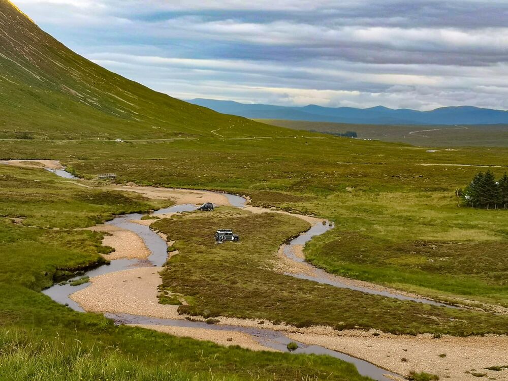 Two 4x4s are parked by a river running through a glen, on the island in the middle surrounded by soft sandy banks. The road is a long way away.