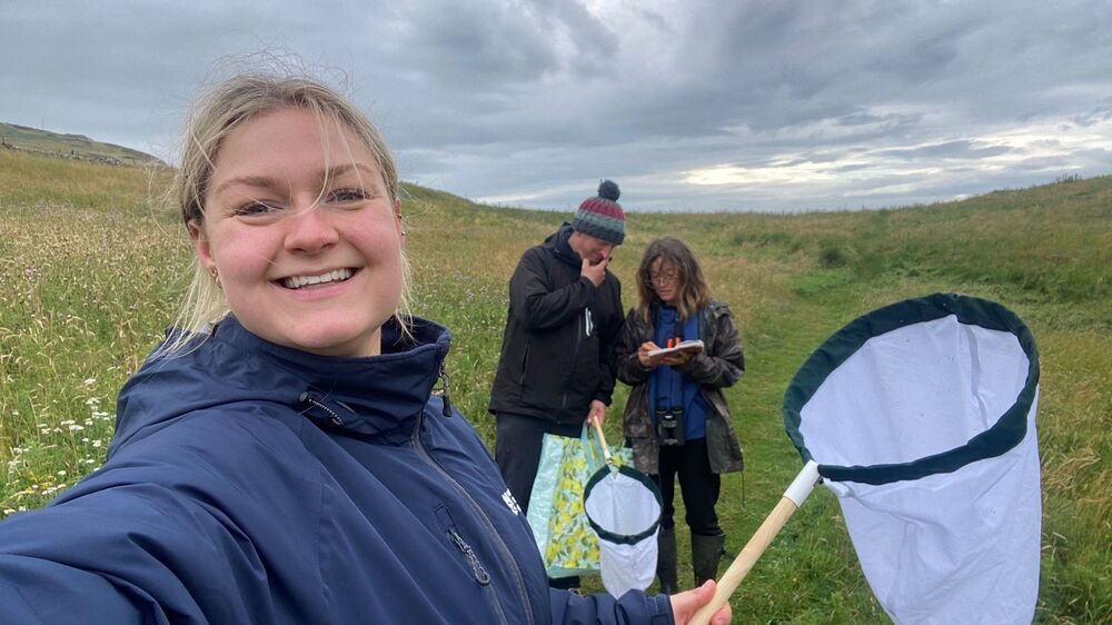 A smiling young woman takes a selfie, as she holds a butterfly net in the other hand. She is surrounded by green hills. Behind her are a man in a woolly hat and dark clothing and a woman in dark clothing, both making notes on a paper pad.