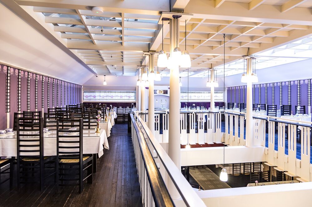 A view from the first floor balcony of the tea rooms, showing Mackintosh chairs arranged around a wooden floor. Long pendant ceiling lights hang down through the balcony space.