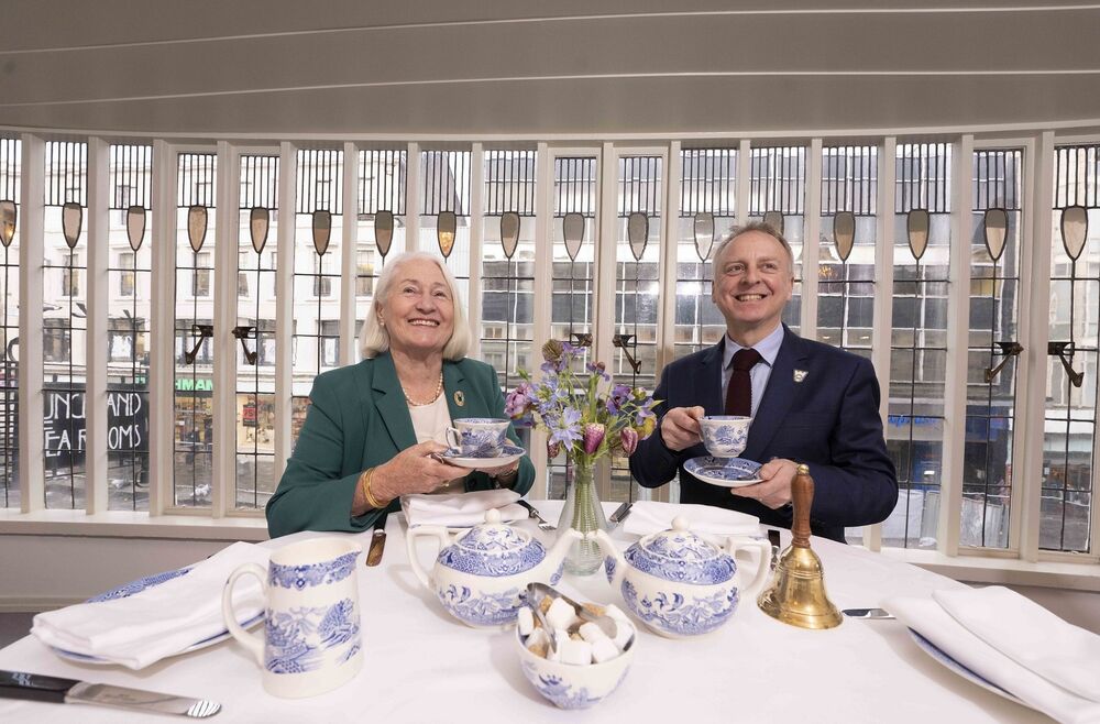 A man and a woman sit at a linen-covered table in a tea room, each holding a blue Willow tea cup and saucer. They are smiling at the photographer.