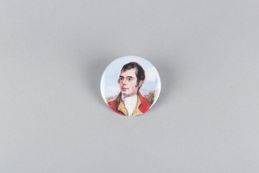 A small round fridge magnet, rather like a button, is displayed against a plain grey background. It features a painting of a young Robert Burns wearing a red jacket.