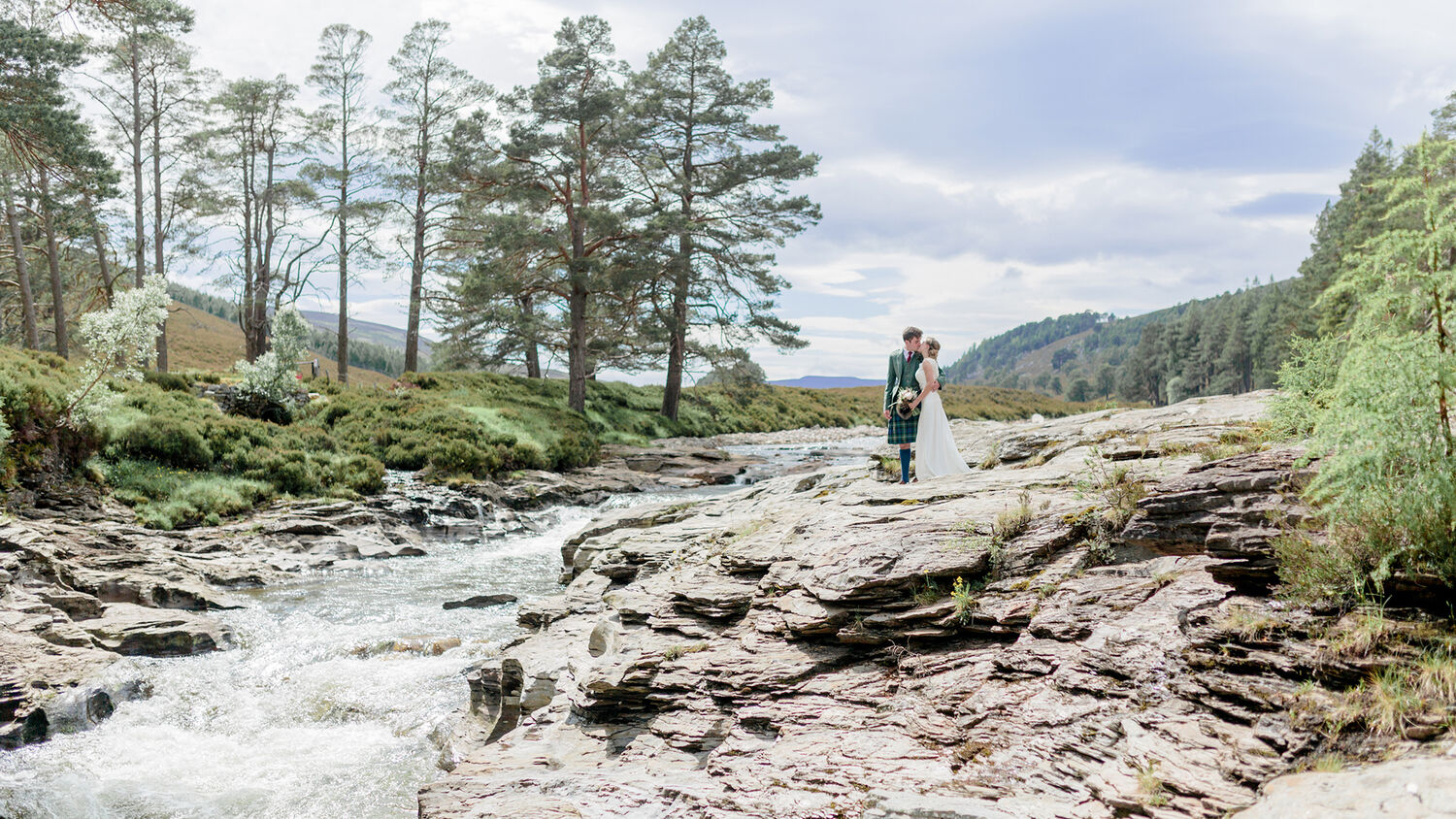 A bride and groom stand on smooth rocks beside a gushing river. Pine trees grow on the far bank and mountains can be seen in the distance.
