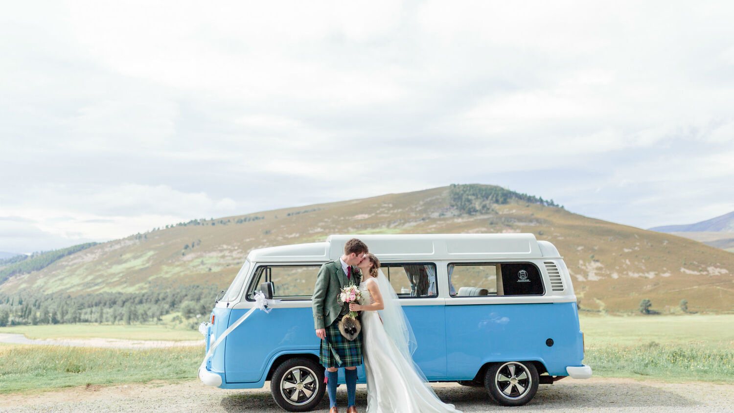 A bride and groom kiss in front of a blue camper van, parked in front of a mountain and moorland.