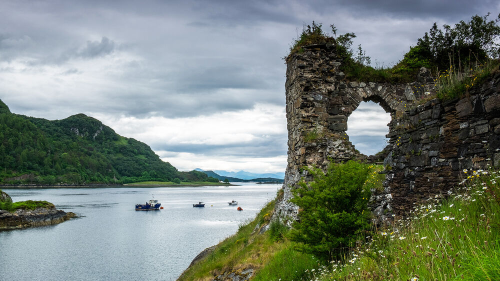A ruined castle perches on a headland with the calm bay behind it. There is a round hole in the old stone wall, which maybe once was a window.