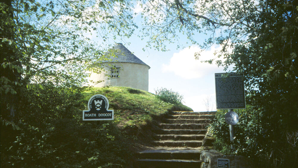 A white round building with a tiled conical roof stands on a hill, at the top of a flight of stairs. Halfway up the stairs in a green, omega-shaped sign in the hillside that says Boath Doocot and has the National Trust for Scotland crest on it.