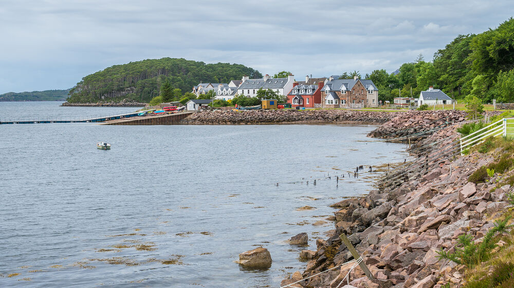 A view from a stony shore that slopes steeply into the water. Along the coastline is a small lumpy island, completely covered in trees. It is very close to the shore. A group of brightly coloured houses stands on the headland directly in front of the island.