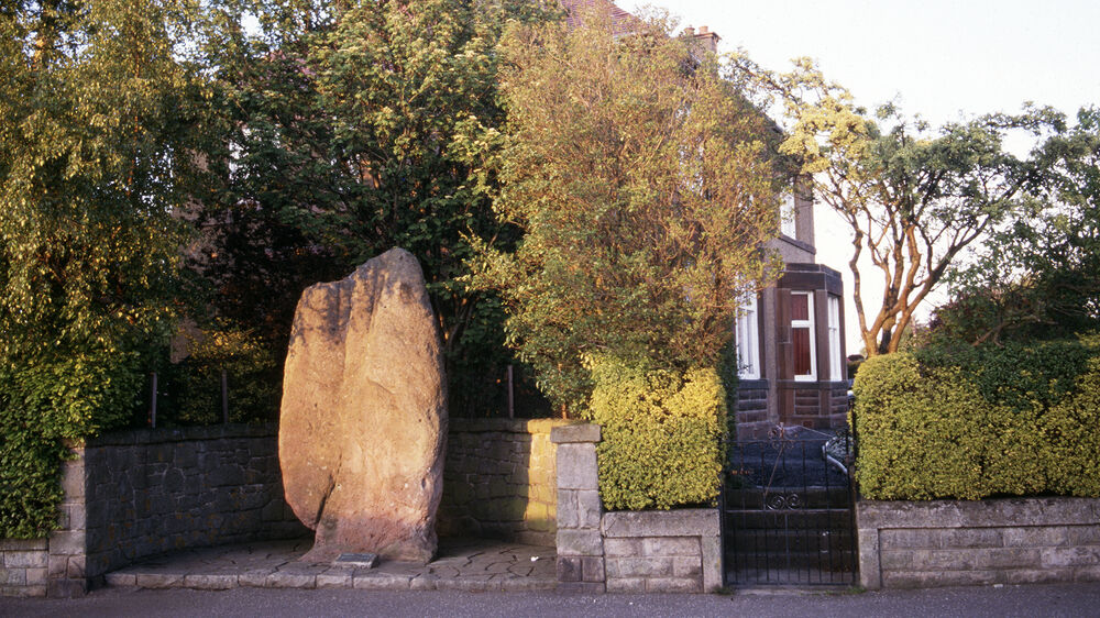 A large and tall granite stone stands in a little alcove at the edge of a residential street. Trees and garden hedges surround it, above a low stone garden wall.