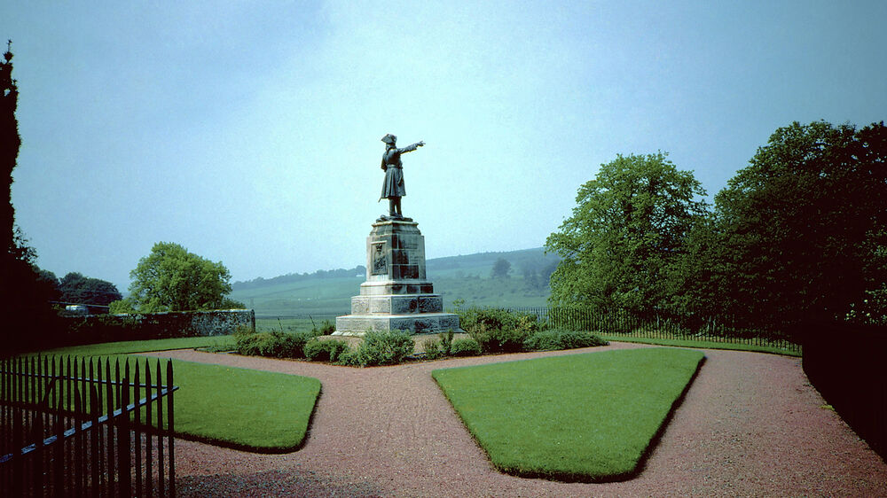 A large stone monument stands in the middle of a park, with neat lawns and gravel paths surrounding it. The white stone plinth is topped with a statue of a man in old military uniform.