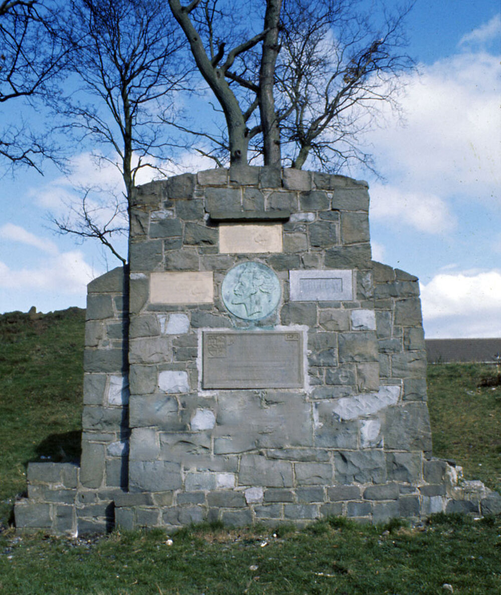A built stone memorial stands on a hillside with a tree behind. There is a circular plaque in the middle of the front wall, surrounded by four rectangular plaques.