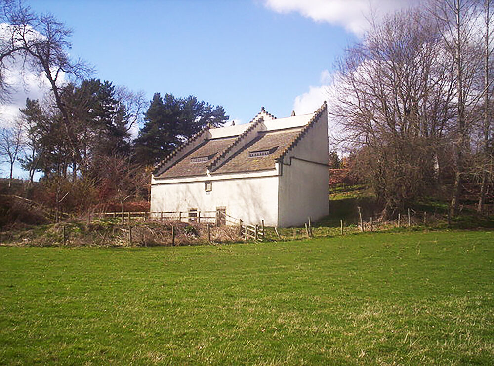 A white stone building with a very large sloping roof stands at the top of a field, with trees either side.
