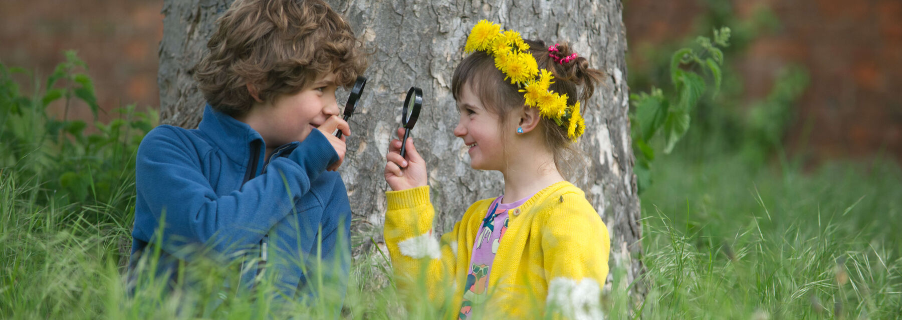 A young boy and girl hold up magnifying glasses whilst sitting underneath a large tree