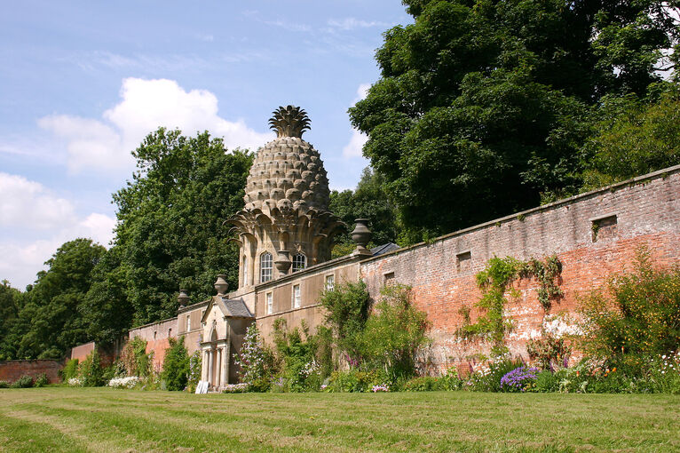 A very tall stone garden wall runs beside a lawn. At the centre of the wall is a stone folly, in the shape of a pineapple. Tall trees stand behind the wall.