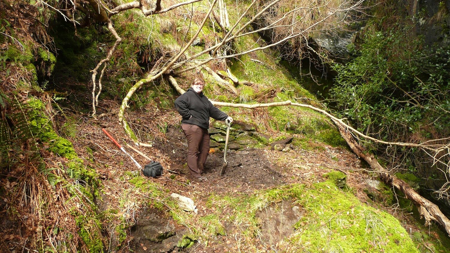 A woman stands in a gorge-like area on a hillside, holding a spade. Branches and tree roots grow horizontally out from the mossy hill, and there appears to be a kind of cave to the left of the woman.