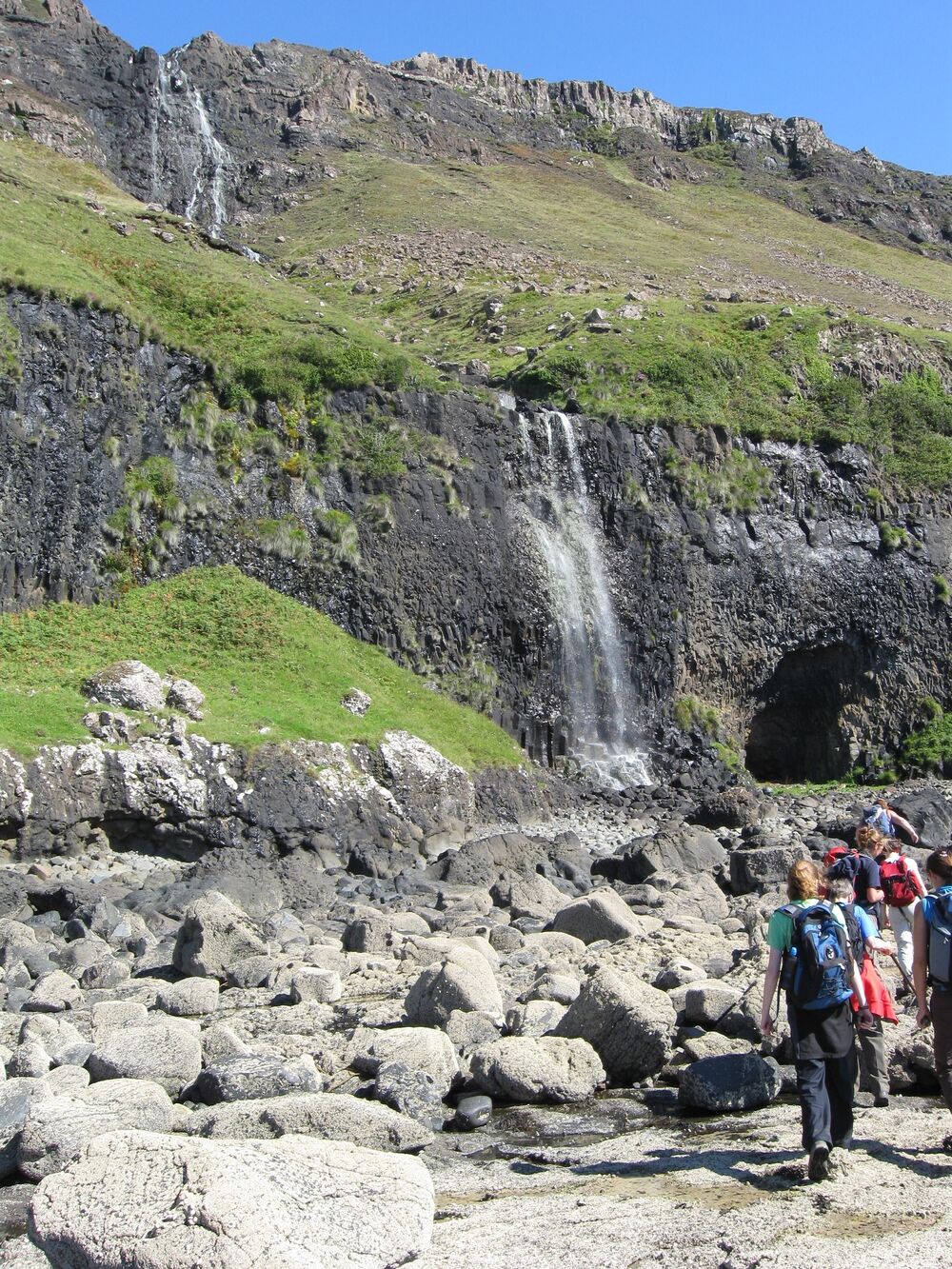 A waterfall tumbles over a cliff to the boulder-strewn beach below. Beside the bottom of the waterfall is a small cave. A group of people walk along the beach towards the cave.