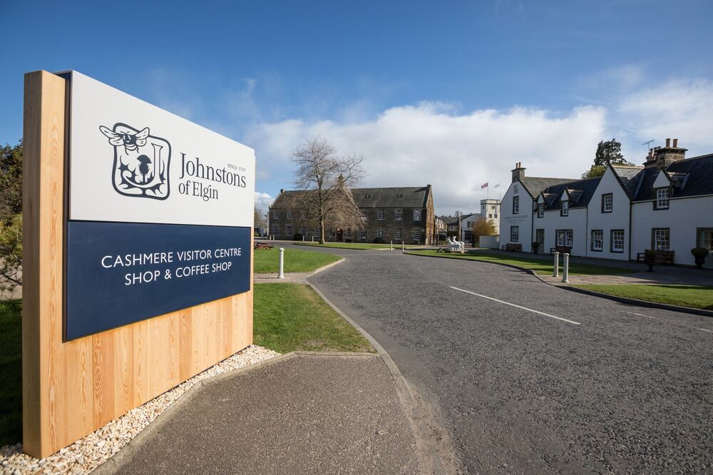 A large sign for Johnstons of Elgin is at the side of a road with buildings in the background.