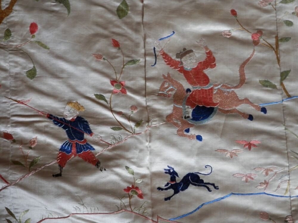 A detail of an embroidered silk panel, showing two Chinese huntsmen. The one on the right is on horseback and wears a red jacket. He is holding a bow in the air. The man on the left is on foot and wears a navy jacket. He is carrying a long spear. A black dog runs between them.