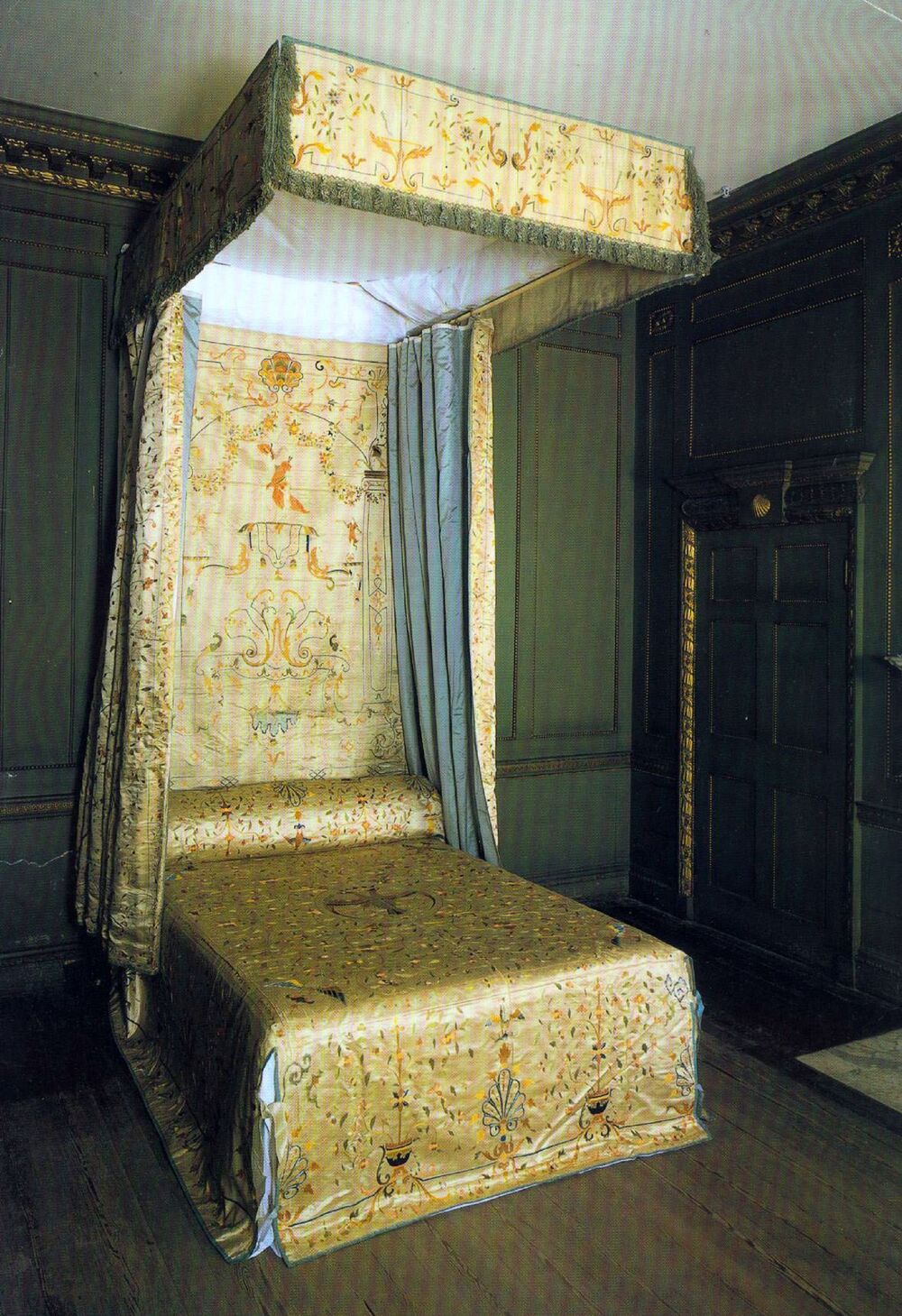 A formal 18th-century-style bed is displayed in a grand bedroom. The bed is covered by exquisite silk panels, featuring various embroidered motifs. The silk hangings are mostly golden in colour and contrast against the darker green wooden panels on the walls.