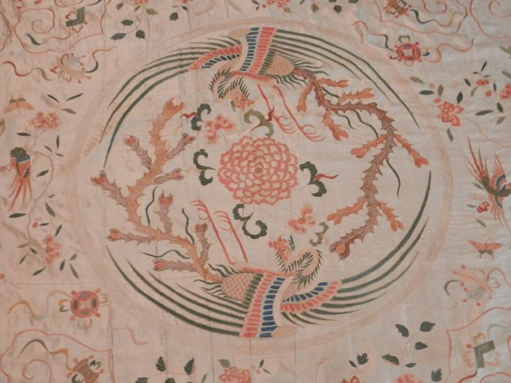 A detail from the centre of an embroidered silk wall panel. It features two phoenixes with fire coming out of their tails, chasing each other round a circle. At the centre of the circle is a red peony motif.