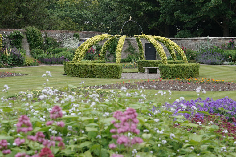 A walled garden, with a closely mown lawn and beds of flowers. There is a low hedge structure in the shape of an open circle with benches inside, with thin bands of hedge also shaped to reach overhead to a centre point.