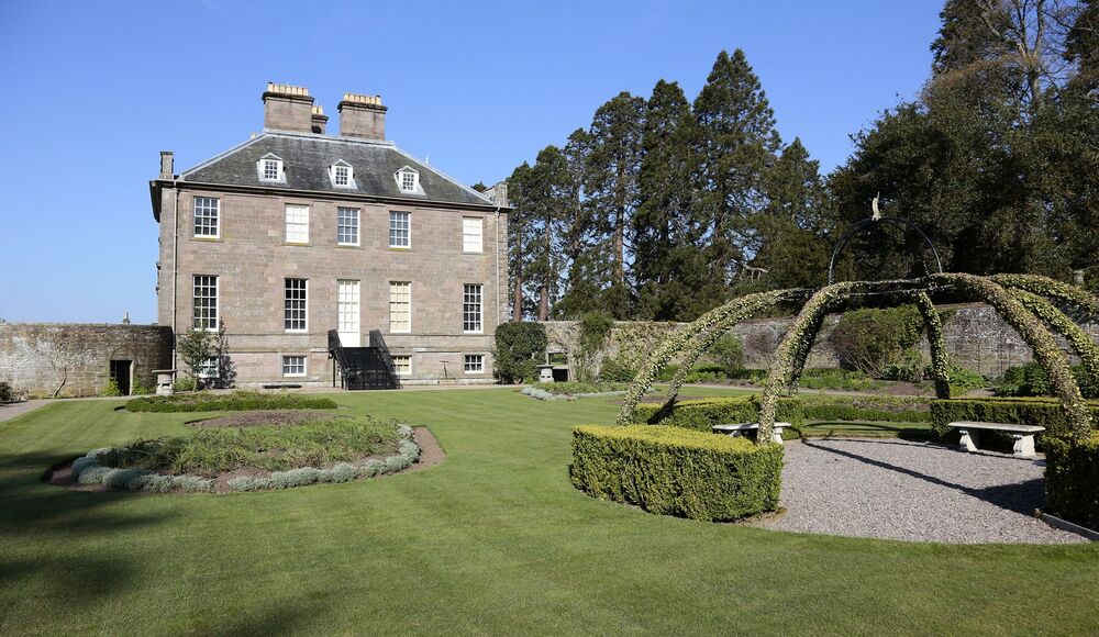 A formal garden lies beside a grand Georgian country house. There are immaculate manicured lawns, with an arched structure covered in climbing roses. Neat box hedges line the paths.