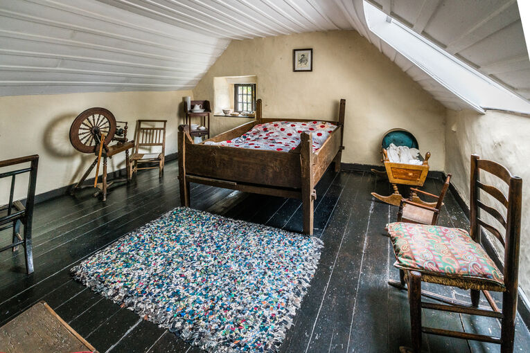 A large double bed and spinning wheel are displayed in an attic room.
