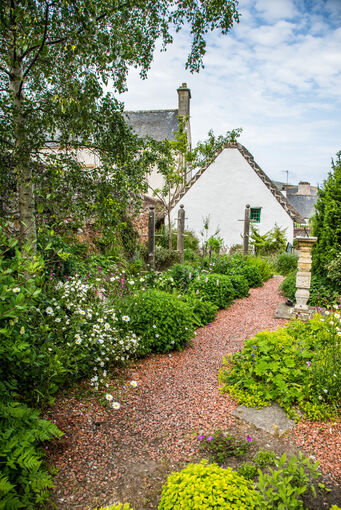 A red gravel path lies between green bushes and flower beds. Hugh Miller’s Cottage can be seen in the background.