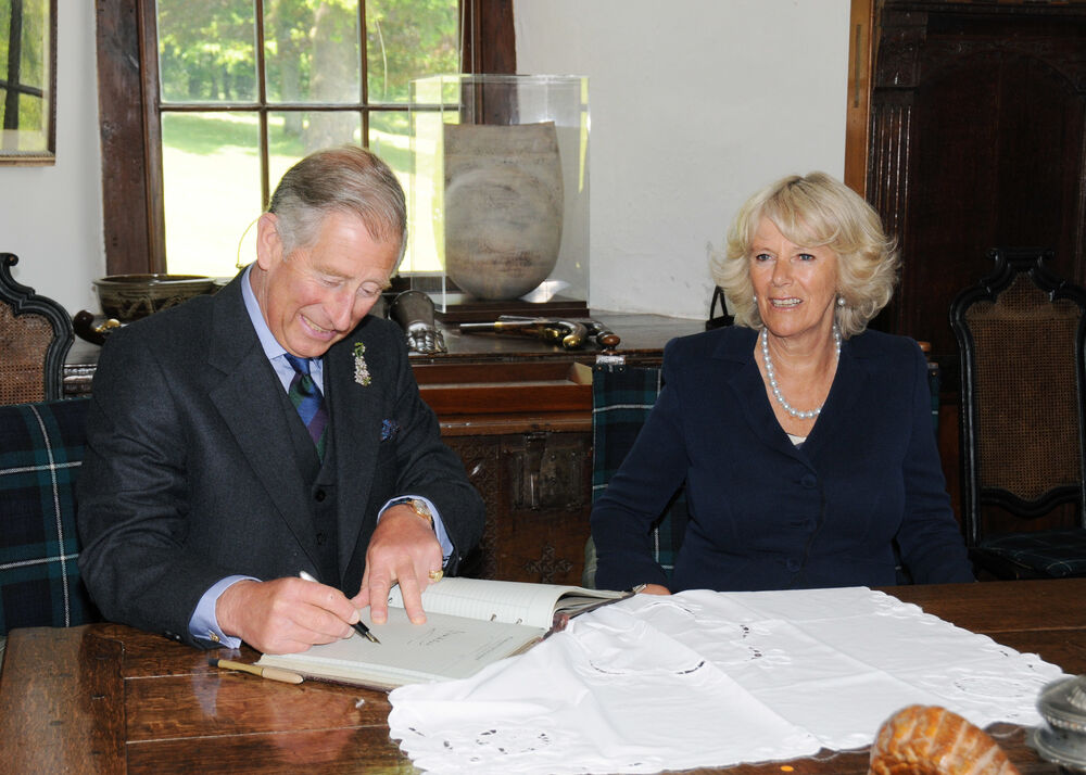 The Duke and Duchess of Rothesay sit at a polished wooden table. A large book is spread out on top of the table. Prince Charles is signing a visitor book.