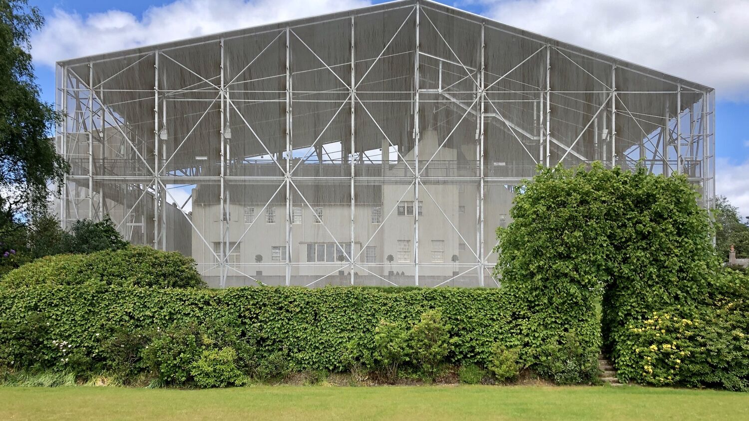 A view of the Hill House, surrounded by a very large metal, chainmail structure. The view is from the garden and it is a sunny day.