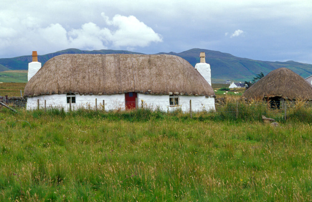 A white thatched cottage stands in a grassy field, with a smaller thatched outbuilding beside. The cottage has a chimney either end, and a small red front door. Mountains can be seen in the background.