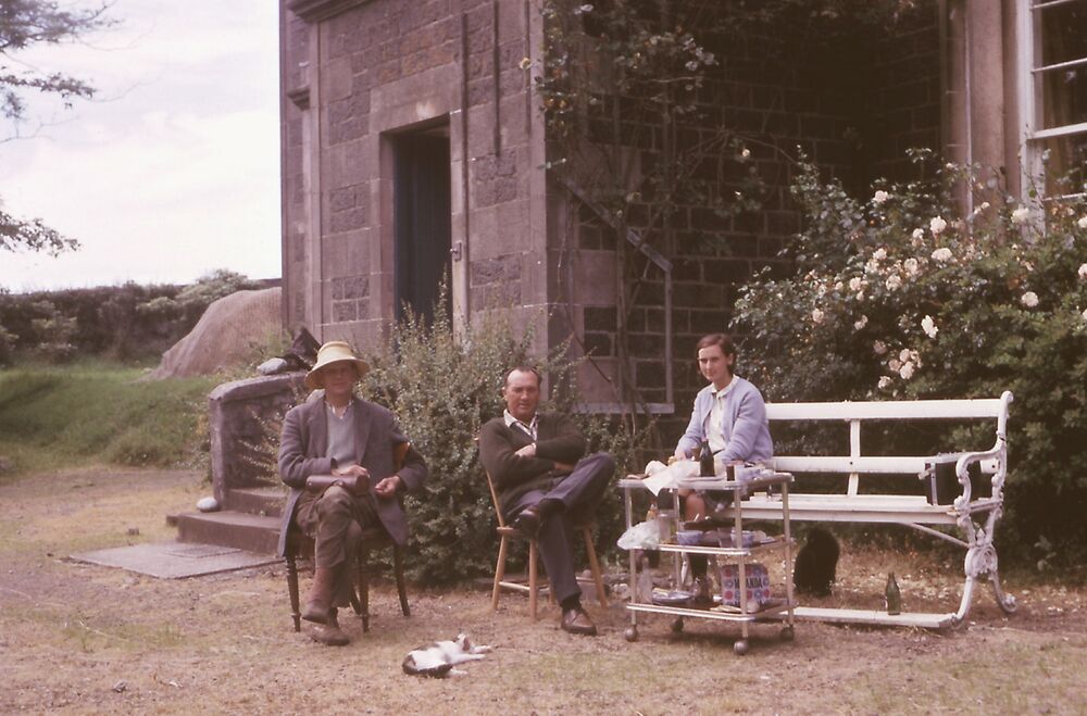Colour photograph of a girl and two men sitting outside a grand stone house.
