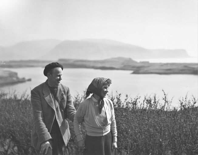 Black and white photograph of a man and woman standing on an island with the sea behind them and another island in the distance.