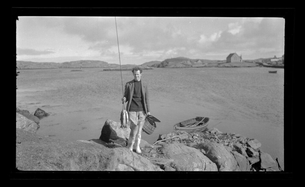 Black and white photograph of a man standing on rocks holding a fishing rod and equipment, with the sea in the background.
