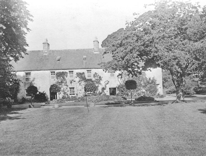 Black and white photograph of a large house with a large area of grass in front of it and a large tree to the right-hand side.