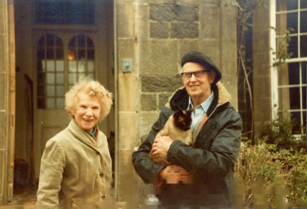 Colour photograph of a man and woman standing outside a house. The man is holding a dog.