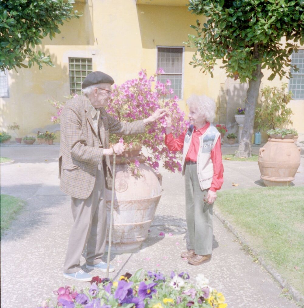 Colour photograph of an elderly man and woman in a garden in front of a large urn filled with flowers.