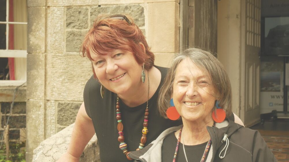 Colour photograph of the head and shoulders of two women outside a stone house.