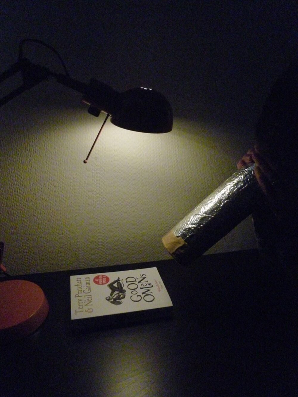 A book on a black table under a lamp, with someone pointing a Pringles tin wrapped in foil (the pinhole camera) at it.