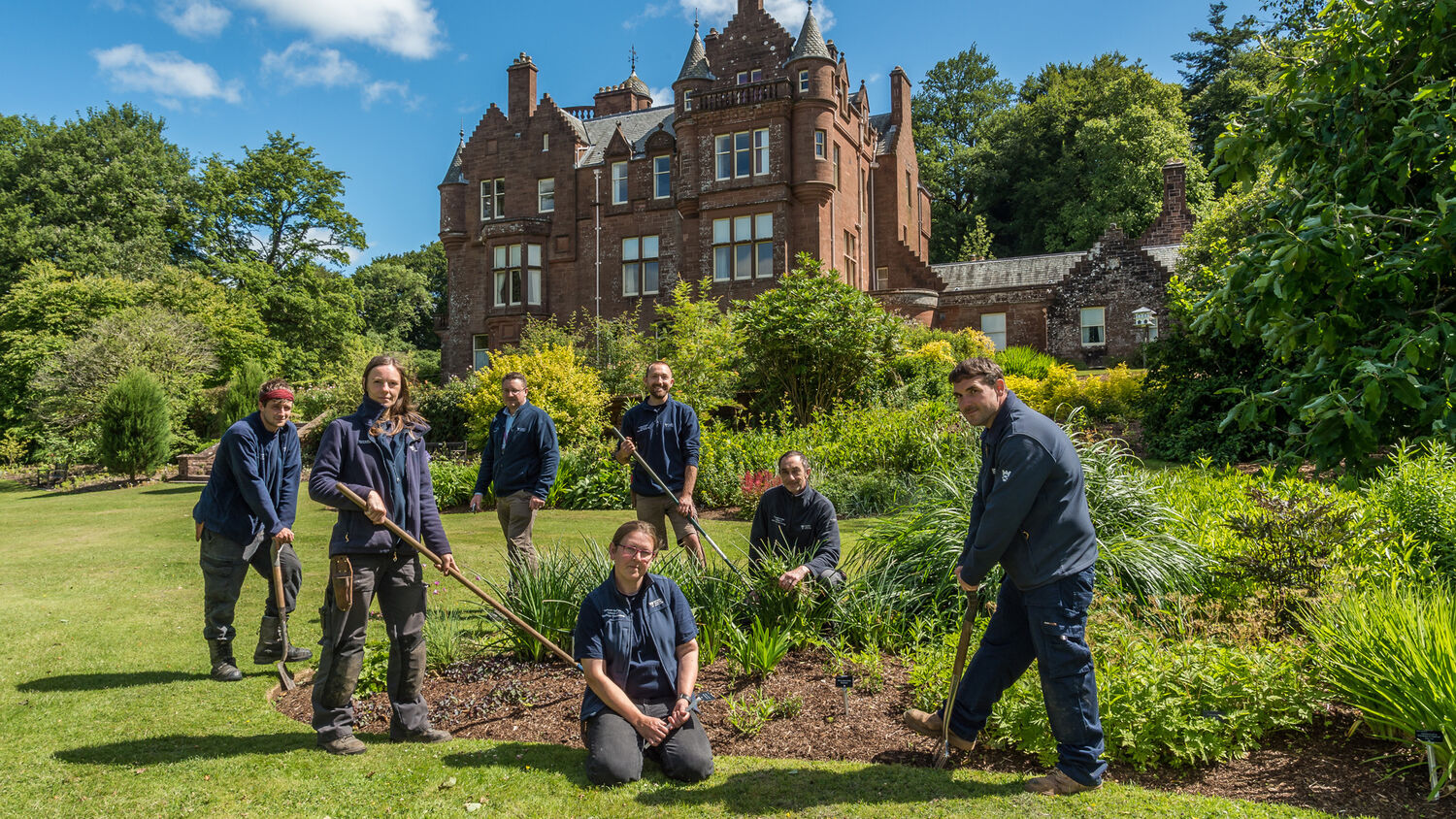 A group of gardeners pose in Threave Garden around a flower bed, with Threave House looming above them! Several are holding hoes or other digging implements.