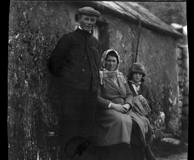 Black and white photograph of three people sitting outside a single-storey stone cottage.