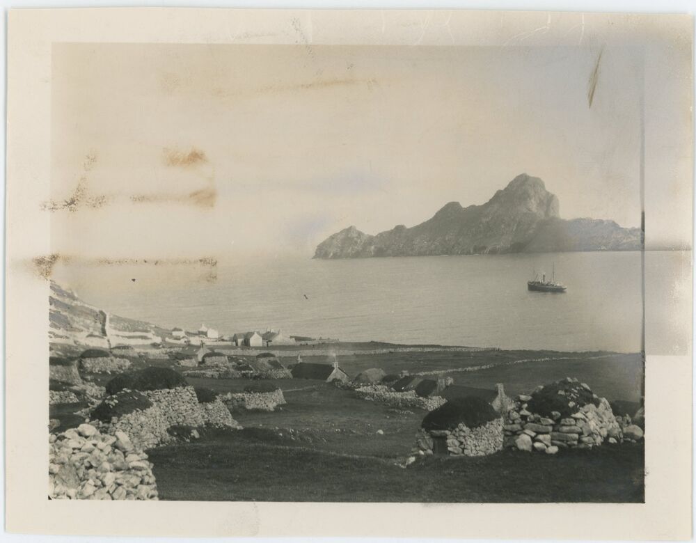 Black and white photograph looking over a bay, with turf-covered cleits in the foreground, and a boat in the bay.