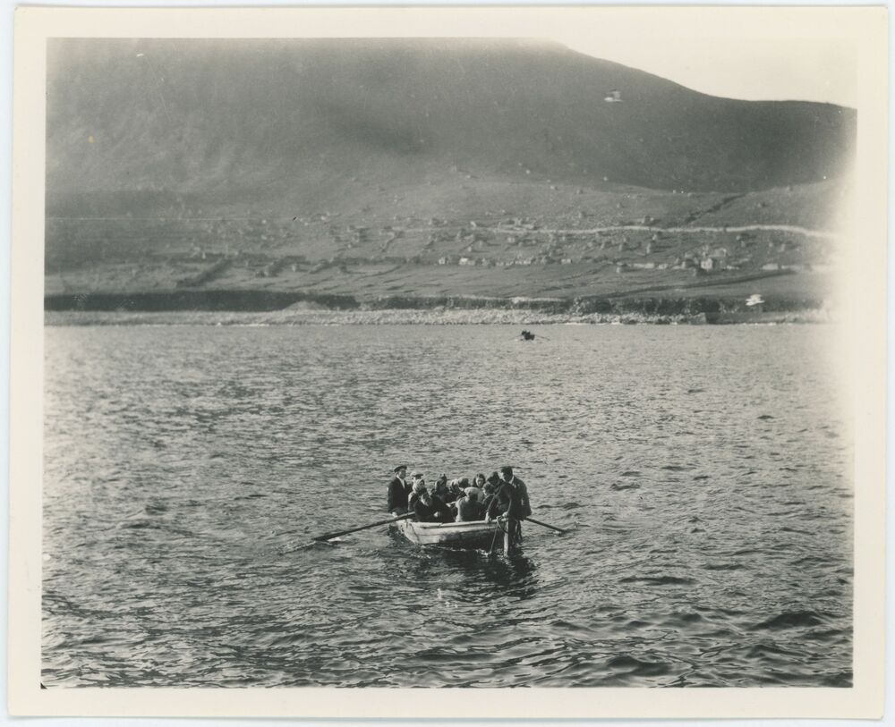 Black and white photograph of a rowing boat full of people in the sea, with an island in the background.