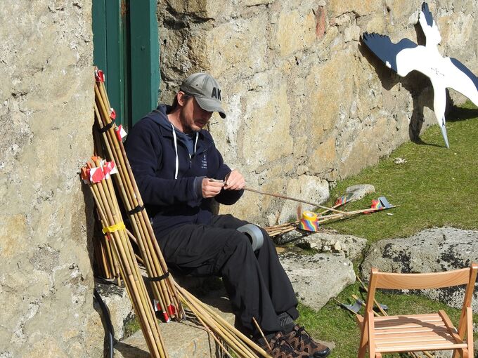 A man sits on the doorstep of a stone cottage on a sunny day, adding bright stickers to bundles of wooden canes. In the background, a model of a gannet rests against the wall.