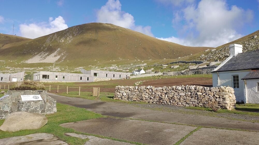 A cluster of new single-storey buildings stand at the foot of a hill, almost completely blending into the landscape. In the foreground is an older stone white cottage with a drystone wall at the front. A tarmac track passes in front of it towards the new buildings.